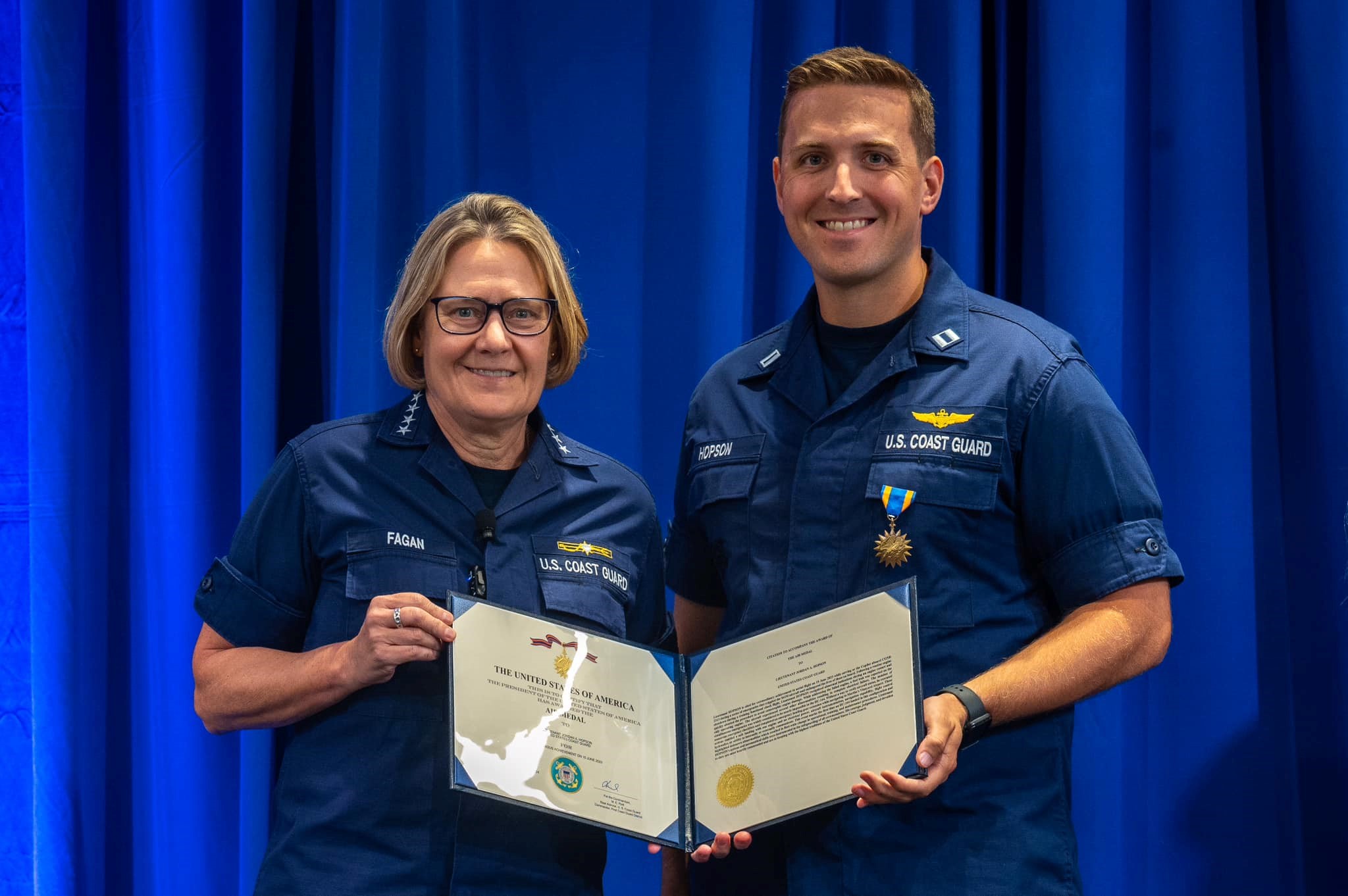 Blue background with Adm. Linda Fagan on the left, Lt. Jordan Hopson on the right, and they are co-holding a decoration. Blue background with Adm. Linda Fagan on the left, Lt. Jordan Hopson on the right, and they are co-holding a decoration.